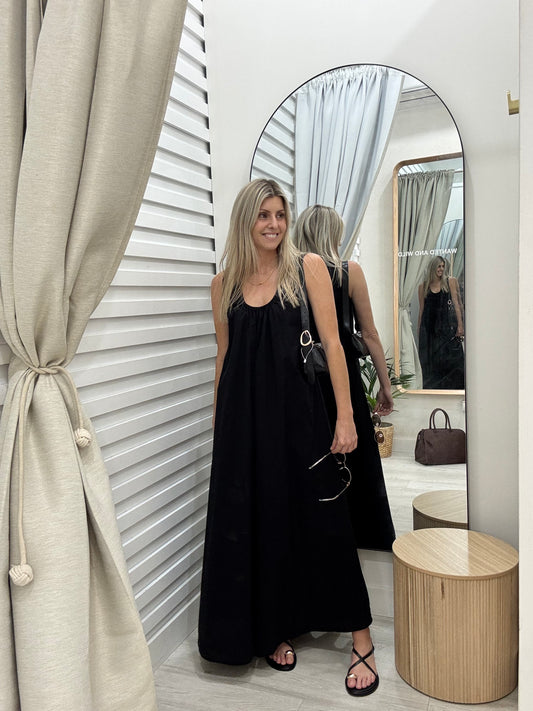 Woman in a black dress standing in front of a mirror with a neutral-colored curtain.