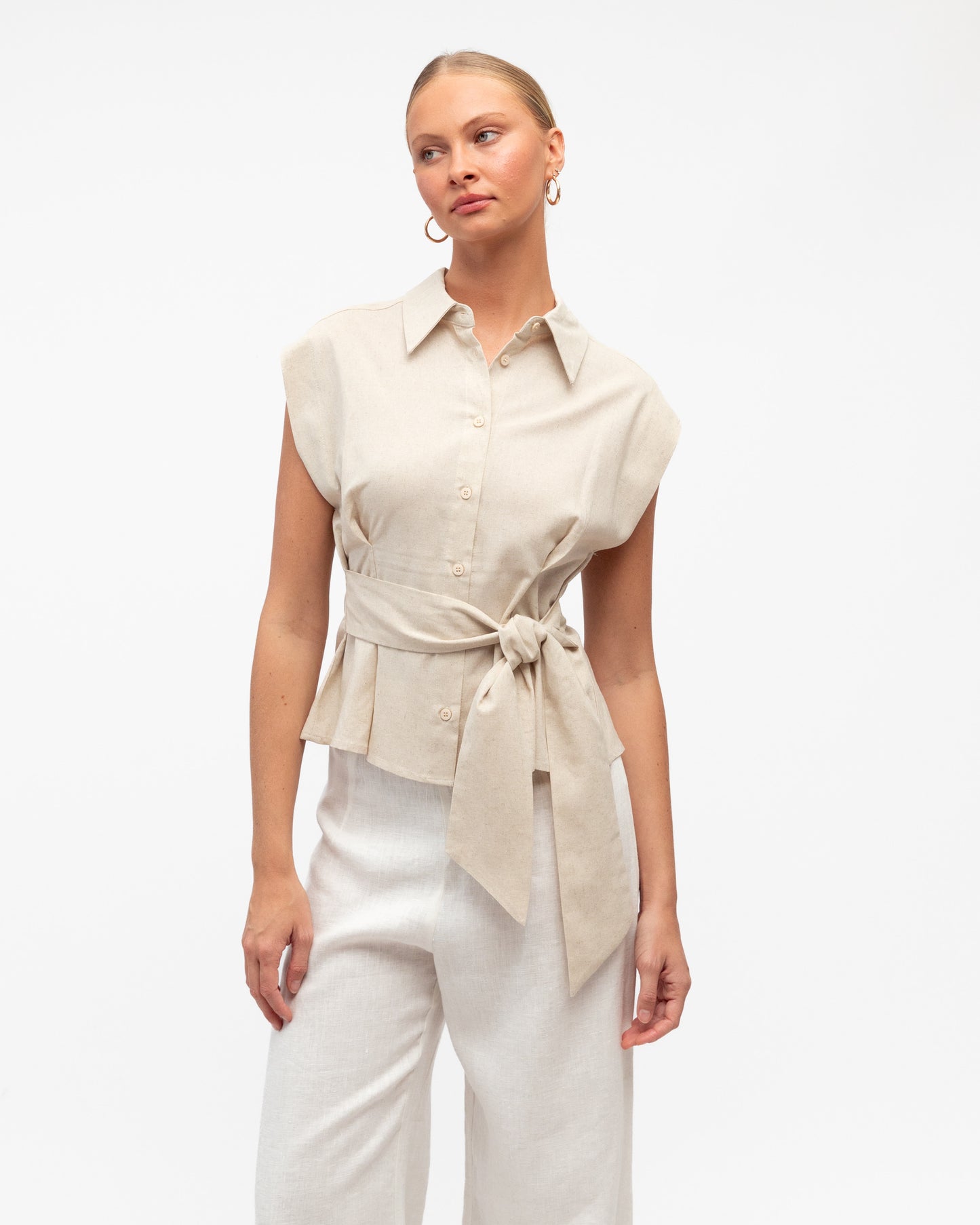 Model wearing a Linen Luxe sleeveless shirt with a tie waist and white pants on a light gray background. White Closet at Wanted and Wild