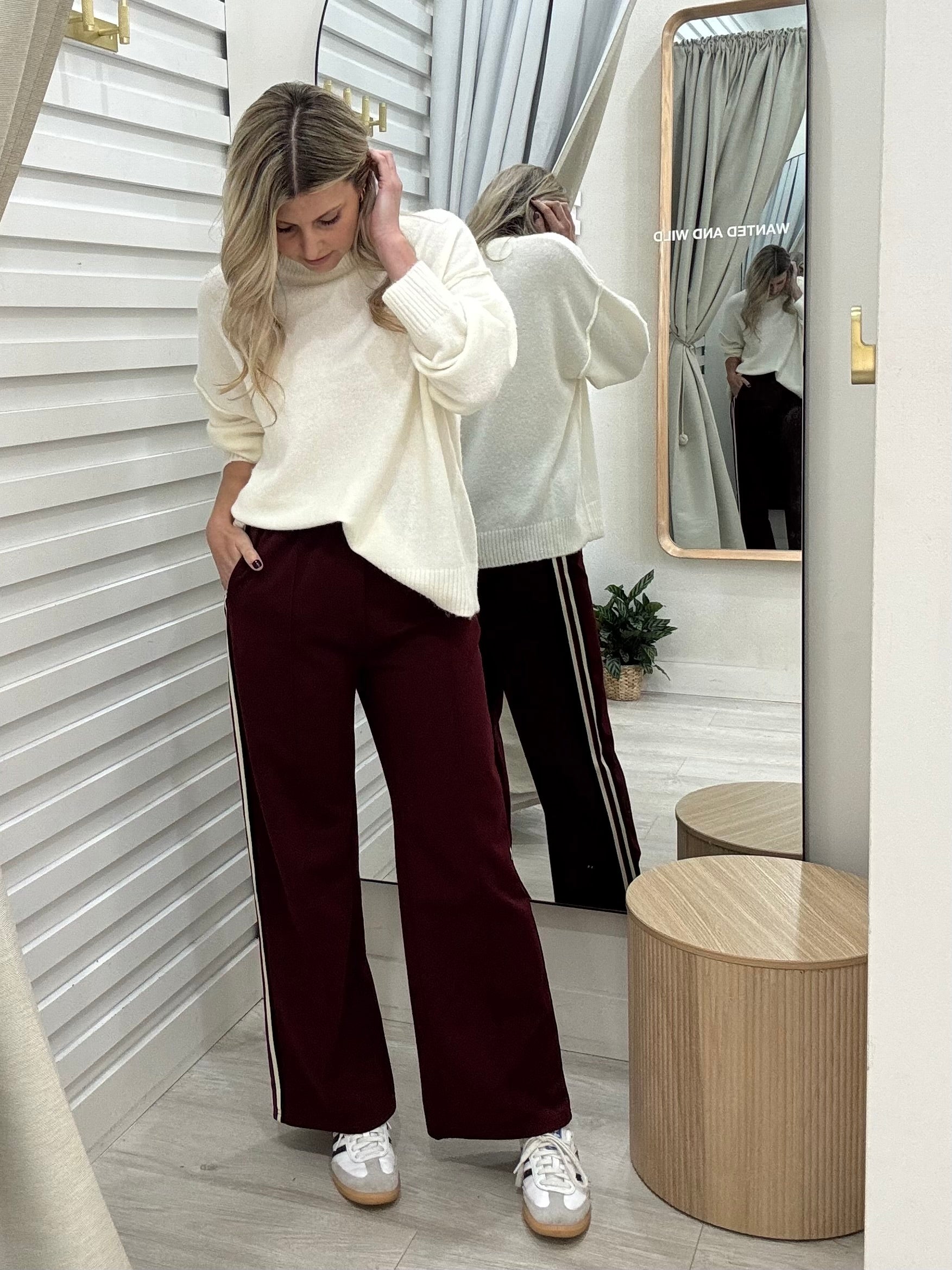 Woman in a cream sweater By Frankie and burgundy pants standing in front of a mirror at Wanted and Wild