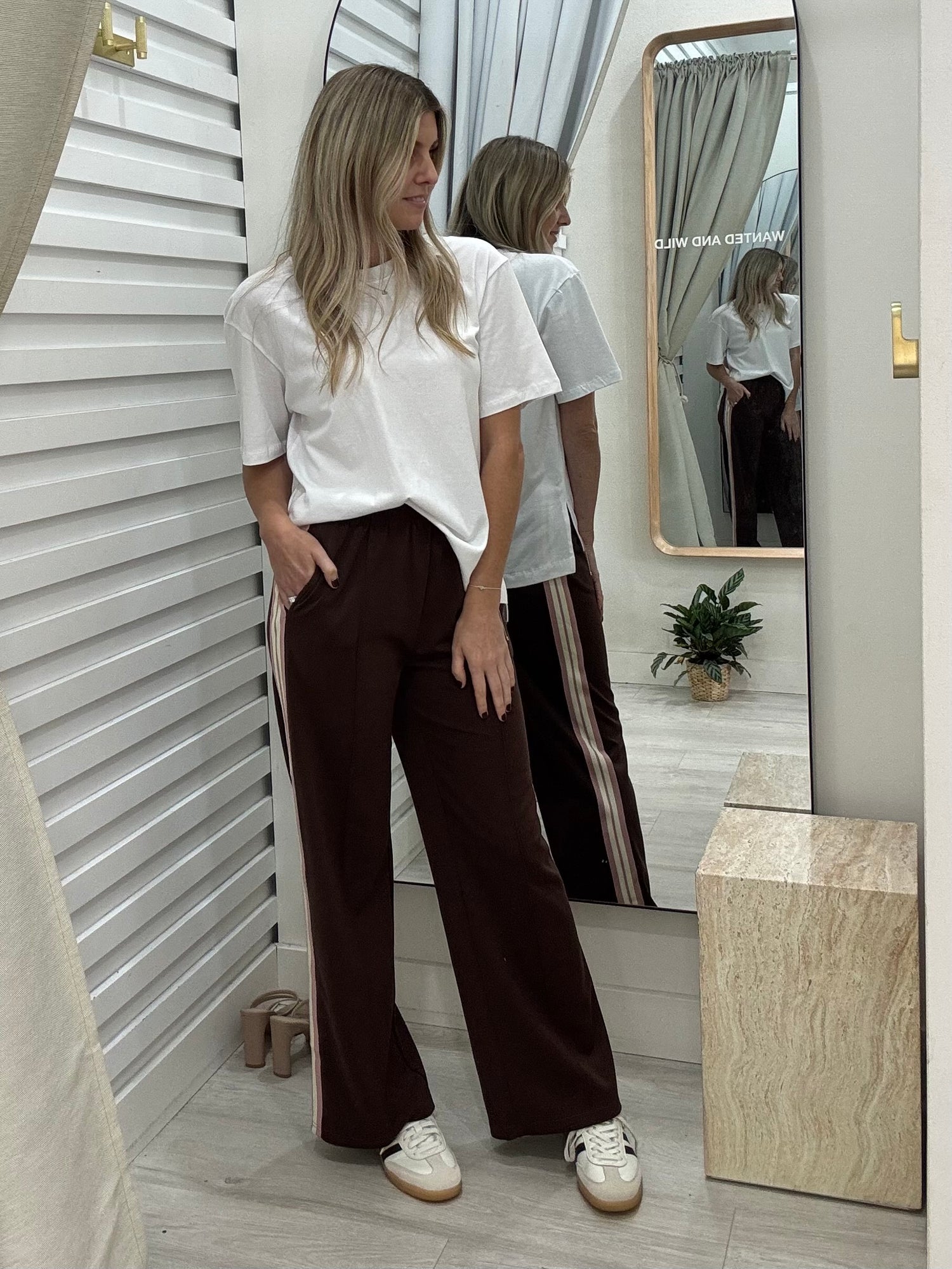 Woman standing in front of a mirror wearing a white t-shirt and brown pants, posing with hands in pockets.