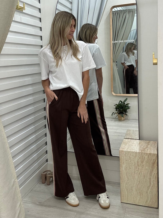 Woman standing in front of a mirror wearing a white t-shirt and brown pants, posing with hands in pockets.