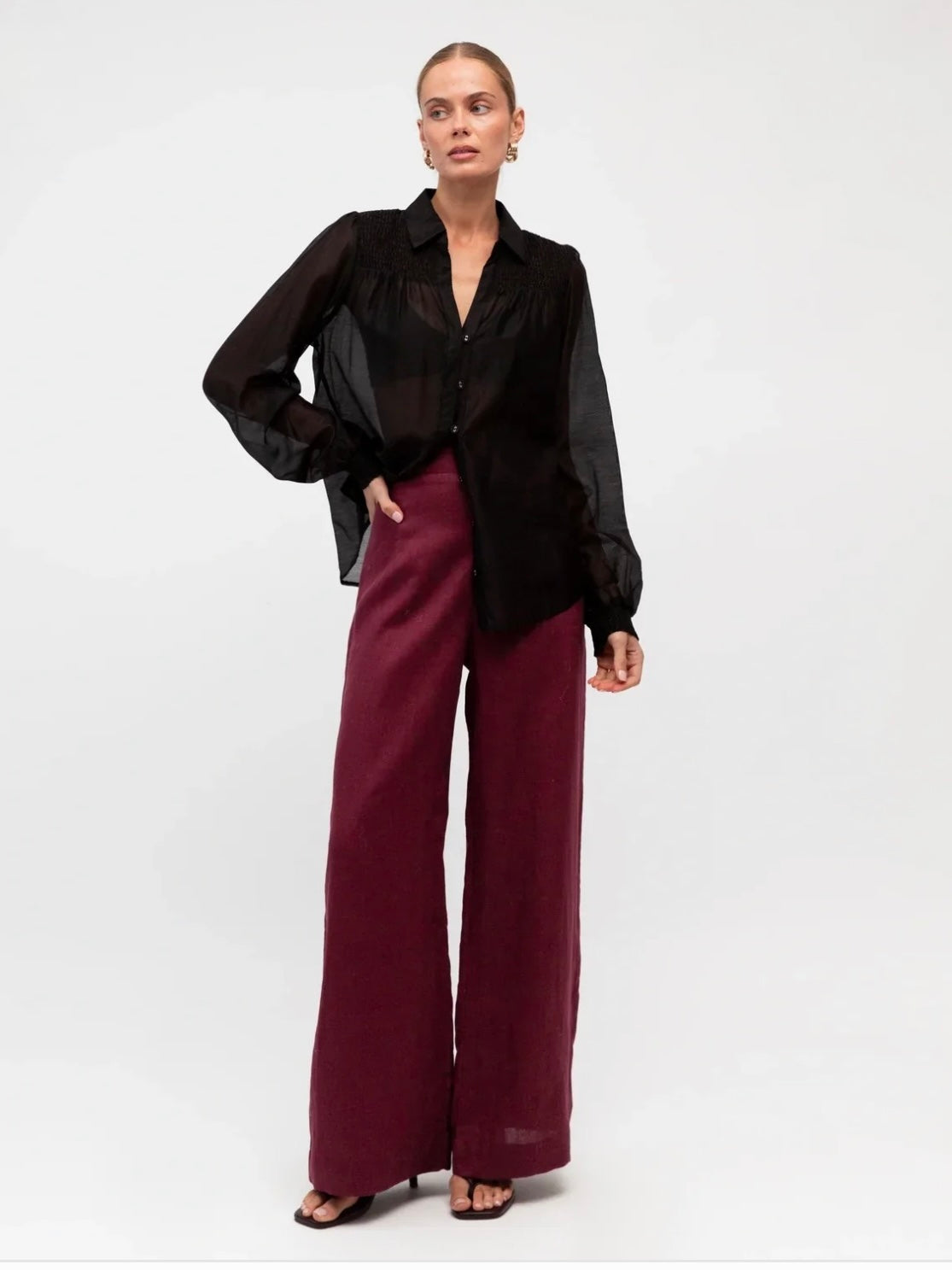 A model wearing a sheer black long-sleeve button-up blouse and high-waisted, wide-leg burgundy trousers, standing against a plain white studio background.