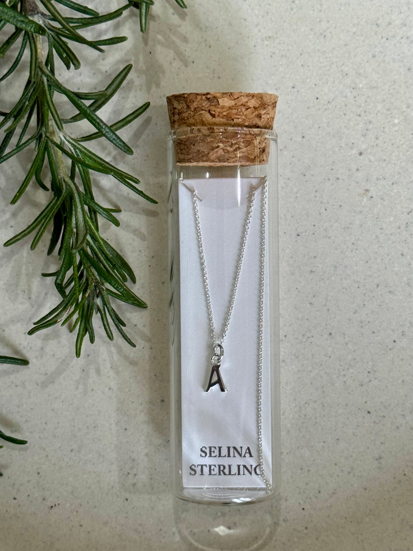 Silver necklace with a letter 'A' pendant in a Selina Sterling packaging on a textured surface with greenery.
