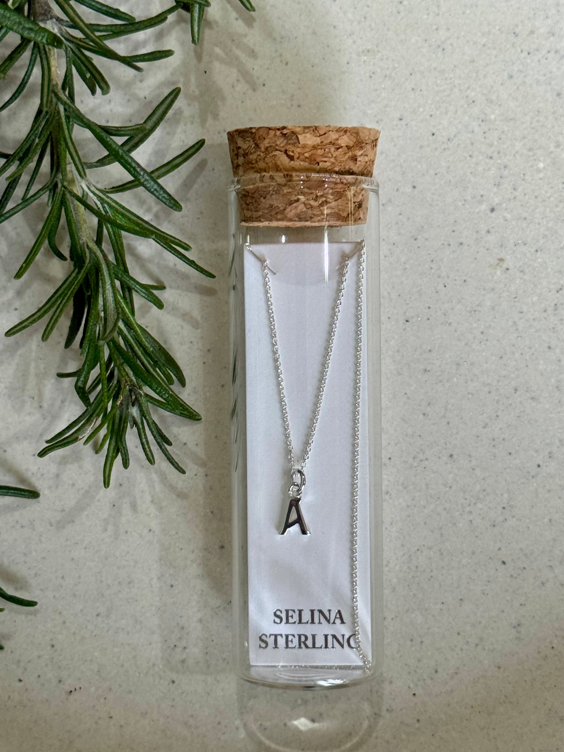 Silver necklace with a letter 'A' pendant in a Selina Sterling packaging on a textured surface with greenery.