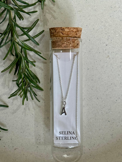 Silver necklace with a letter 'A' pendant in a Selina Sterling packaging on a textured surface with greenery.
