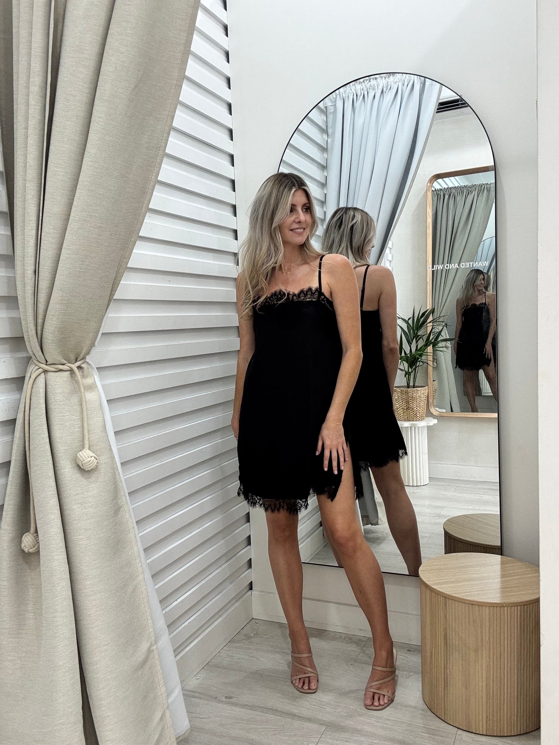 Woman in The Ace dress in black, standing in front of a mirror  at Wanted and Wild. Wearing Brooklyn heels in nude at Wanted and Wild.