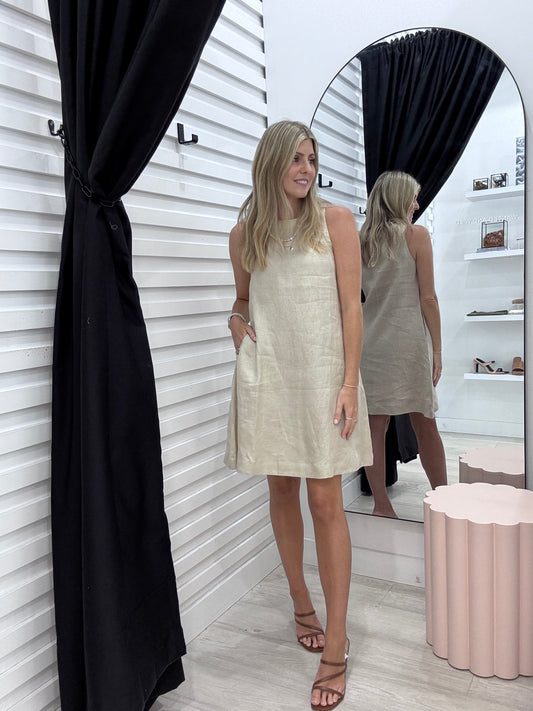 A woman standing in Wanted and Wild wearing the Atlas sleeveless, oatmeal-colored linen knee-length dress with a round neckline, 