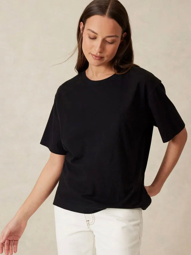 A woman wearing a black short-sleeve crewneck t-shirt and white pants, standing against a neutral beige textured wall.