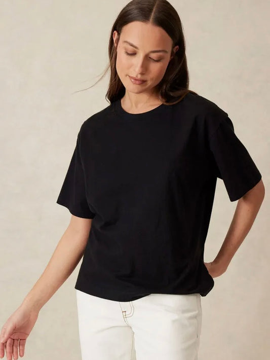 A woman wearing a black short-sleeve crewneck t-shirt and white pants, standing against a neutral beige textured wall.