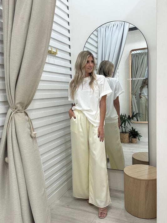 Woman in a white top and beige pants standing in front of a mirror in a room with neutral decor.